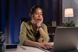 asiatico donna uomo d'affari Lavorando su sua il computer portatile per preparare per il presentazione Domani mattina. lei guardato così assonnato e stanco quello lei sbadigliato molto in ritardo notte a casa. foto