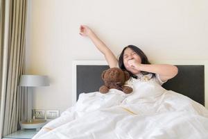 ritratto di asiatico ragazza risveglio, scia su su il letto. carino asiatico ragazza allungamento sua braccia. foto