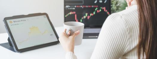 investitore analizzando scambio mercato, asiatico giovane attività commerciale donna mano Tenere caffè tazza, commerciante su grafico grafici su il computer portatile computer, tavoletta per commercio scorta, guardare a schermo con diagrammi a casa. foto