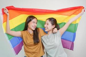 contento lesbica, bellissimo asiatico giovane Due donne, ragazza gay, coppia amore momento la spesa bene tempo insieme, Tenere o agitando lgbt arcobaleno, orgoglio bandiera su isolato bianca parete sfondo insieme a casa. foto