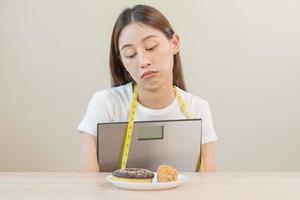 dieta, attraente asiatico giovane donna, ragazza trattenuto per mangiare ciambella, forno e fritte pollo, veloce cibo per perdere, perdita il peso, abbracciare peso bilancia su tavolo a casa. passione, tentazione quando Affamato. foto