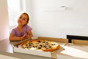 affascinante contento giovane ragazza ridere e pungente via grande fetta di fresco fatto Pizza. lei sedersi a bianca sedia nel provence stile interno, Sorridi e godere soleggiato giorno e delizioso pasto. lei ha lungo bionda capelli. foto