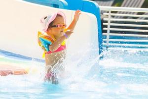 contento ridendo poco ragazza giocando su acqua diapositiva nel all'aperto nuoto piscina su caldo estate giorno. bambini imparare per nuotare. bambino indossare sole protezione eruzione cutanea guardia scorrevole su acqua terreno di gioco nel tropicale ricorrere foto