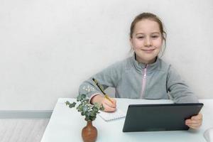 carino poco ragazza seduta a il tavolo scrittura giù qualcosa a partire dal il tavoletta, in linea apprendimento foto