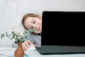 carino poco ragazza seduta a il tavolo sorridente sbirciando su a partire dal dietro a un Aperto computer portatile, in linea apprendimento foto