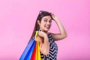 immagine di un' bellissimo giovane brunetta donna nel estate vestito indossare occhiali da sole in posa con shopping borse e guardare a telecamera al di sopra di rosa sfondo. foto