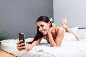 bellissimo giovane sorridente brunetta donna con oro cosmetici maschera, occhio cerotti dire bugie nel bianca letto e utilizzando un' Telefono nel sua Camera da letto. attraente donna è messaggistica su telefono foto