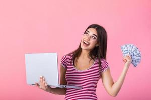 ritratto di un eccitato soddisfatto ragazza Tenere i soldi banconote e il computer portatile computer. contento donna con fortunato i soldi. foto di bellissimo donna con computer portatile, isolato su rosa sfondo