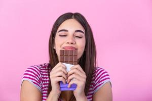bello sorridente adolescenziale ragazza mangiare cioccolato. Immagine di contento carino giovane donna in piedi isolato al di sopra di rosa sfondo mangiare cioccolato. foto