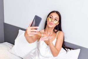 giovane contento donna con oro cosmetici maschera, occhio cerotti utilizzando mobile Telefono per video chiamata , Esprimere a gesti baci per gli amici. bellissimo ragazza fabbricazione FaceTime video chiamata con smartphone dire bugie su il letto. foto