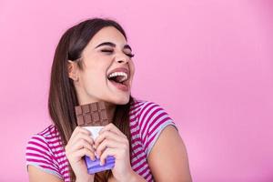 contento giovane bellissimo signora mangiare cioccolato e sorridente. ragazza degustazione dolce cioccolato. giovane donna con naturale rendere su avendo divertimento e mangiare cioccolato isolato su rosa sfondo foto