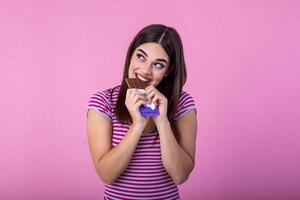 bello sorridente adolescenziale ragazza mangiare cioccolato. Immagine di contento carino giovane donna in piedi isolato al di sopra di rosa sfondo mangiare cioccolato. foto