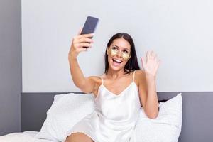 sexy bellissimo donna con oro cosmetici maschera, occhio cerotti con un' smartphone nel sua letto. bellissimo giovane donna nel seta camicia da notte lettura testo Messaggio su sua mobile Telefono e sorridente. foto