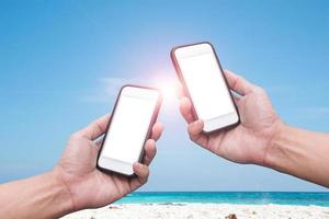 mano che tiene uno smartphone su una spiaggia e uno sfondo di cielo blu foto