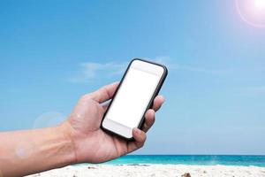 mano che tiene uno smartphone su una spiaggia e uno sfondo di cielo blu foto