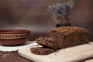 nero pane con cereali affettato su un' di legno taglio tavola contro un' Marrone sfondo. Il prossimo per il pane è un' argilla piatto, forchetta, cucchiaio e bianca cotone tovaglia foto