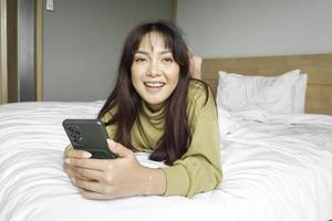 un' sorridente giovane asiatico donna è ridendo mentre Tenere sua Telefono e dire bugie su il letto foto