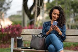 giovane africano americano donna d'affari sorridente e guardare a sua smartphone mentre controllo sociale media nel un' pubblico parco foto