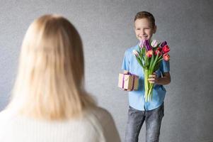 carino ragazzo nel un' blu camicia dà un' mazzo di tulipani e un' scatola con un' regalo per mamma foto