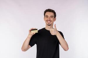 ritratto di giovane sorridente bello uomo nel casuale Abiti mostrando credito carta e pollice su isolato al di sopra di bianca sfondo foto