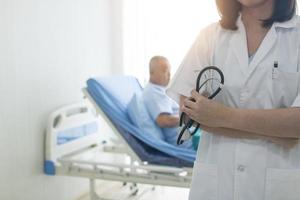 medico con le braccia incrociate tenendo lo stetoscopio con offuscata paziente sul letto in background foto