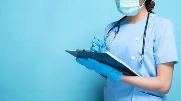 medico che indossa maschera facciale, stetoscopio e guanti blu che scrivono negli appunti su uno sfondo blu foto