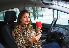 donna d'affari nel un' pelliccia cappotto con rosso labbra invio un' testo Messaggio e potabile caffè mentre guida foto