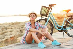 spensierato donna con bicicletta seduta su un' di legno sentiero a il mare foto