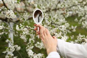 all'aperto primo piano della giovane mano femminile che tiene un piccolo specchio retrò nel giardino fiorito il giorno di primavera. modella guardando in un piccolo specchio, in posa in strada, vicino ad alberi in fiore. concetto di moda femminile foto