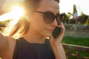 donna nel occhiali da sole parlando su il smartphone mentre a piedi giù il strada a tramonto, vicino su foto
