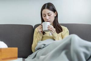 malato, male o dolore asiatico giovane donna, ragazza dolorante gola con bicchiere, boccale di caldo acqua, mal di testa avere un' febbre, influenza nel debolezza, seduta rilassato su divano letto a casa. Salute cura persona su virus di stagione. foto