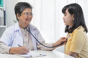 maturo asiatico medico psichiatra, generale donna medico consulenza ha riguardato con adolescenziale ragazza, bambino paziente, utilizzando stetoscopio per ascoltare, appuntamento a clinica. Salute cura, dai un'occhiata su medico. foto