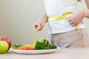 sottile forma asiatico giovane donna, ragazza in piedi, mano nel misurare nastro in giro vita, corpo sua avere un' verdure su tavolo come bianca sfondo. dieta sessione per benessere Salute, mangiare bene cibo le persone. foto