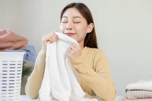 sentire morbidezza, sorridente asiatico giovane donna, ragazza toccante soffice asciugamano cotone, odorare fresco pulito Abiti su tavolo dopo lavaggio, lavanderia, asciutto. domestico Lavorando a casa. lavanderia e domestica. foto