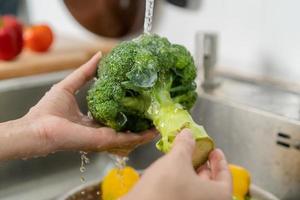 vicino su asiatico giovane donna lavaggio broccoli, pomodoro, carota fresco la verdura, paprica con spruzzo acqua nel bacino di acqua su Lavello nel cucina, preparazione fresco insalata, cucinando pasto. salutare cibo le persone. foto