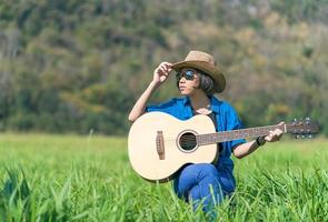 donne corto capelli indossare cappello e occhiali da sole sedersi giocando chitarra nel erba campo foto