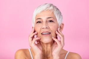 ritratto di bellissimo anziano donna toccante sua Perfetto pelle e guardare a telecamera. avvicinamento viso di maturo donna con rughe massaggio viso isolato al di sopra di rosa sfondo. invecchiamento processi concetto. foto