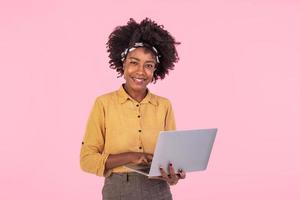 africano americano donna Tenere il computer portatile sorridente e guardare a telecamera. giovane bellissimo donna sensazione felice, in piedi isolato al di sopra di rosa sfondo foto