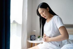 asiatico donna infelice soffre a partire dal stomaco dolore nel il Camera da letto, lei avendo mal di stomaco perché di mestruazione o cronico gastrite. foto