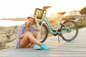 spensierato bella donna con bicicletta seduta su un' di legno sentiero a il mare, avendo divertimento e rilassante foto