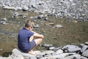 poco ragazzo seduta su rocce con il suo indietro guardare a il montagna lago. foto