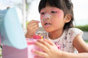 adorabile poco bambino asiatico ragazza vernici sua bocca con rosa bambini teste e sembra nel il specchio. un' bambino giochi a casa nel un' giocattolo bellezza salone. aumentare apprendimento sviluppo per bambini in età prescolare. foto