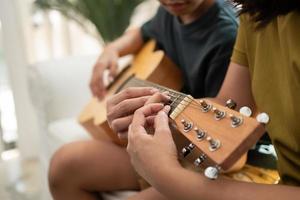 asiatico ragazzo giocando chitarra con mamma nel il vivente camera per insegnamento lui figlio giocare chitarra, sentire apprezzato e incoraggiato. concetto di un' contento famiglia, apprendimento e divertimento stile di vita, amore famiglia cravatte foto