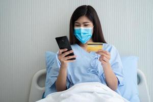 contento asiatico donna indossare un' medico maschera e Tenere finto su credito assicurazione carta e smartphone nel un' ospedale letto per dai un'occhiata Salute assicurazione eleggibilità. assicurazione politica di banca, pagamento medico foto