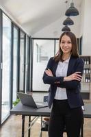 un' elegante bianca donna d'affari nel un' nero completo da uomo in piedi dentro il ufficio nel mani con un' mobile Telefono, tavoletta, e il computer portatile in attesa per i clienti per contatto, attività commerciale persone concetto. foto