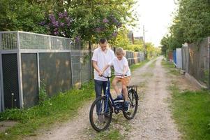 un' carino ragazzo nel un' bianca maglietta impara per cavalcata un' bicicletta con il suo papà e ride. papà insegna il suo figlio. foto