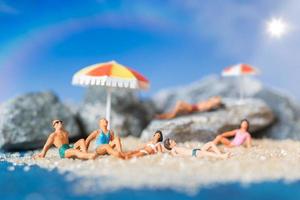 persone in miniatura che indossano costumi da bagno rilassante sulla spiaggia con uno sfondo blu, concetto di estate foto