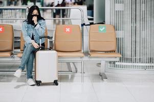 donna viaggiatore che indossa la maschera per il viso tosse mentre è seduto sulla sedia di allontanamento sociale foto