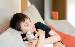 ritratto ragazzo seduta su divano con cane giocattolo Guardando tv, giovane ragazzo seduta su divano guardare su in profondità nel pensiero. positivo bambino rilassante a casa su fine settimana, bambini stile di vita concetto foto