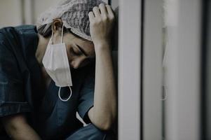 stanco depresso donna asiatica scrub infermiera indossa maschera facciale uniforme blu siede sul pavimento dell'ospedale, giovane donna medico stressato dal duro lavoro foto